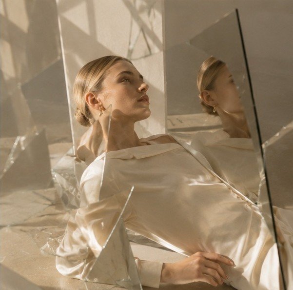 A woman in ivory silk reclining among angled mirror fragments, her reflection repeated in the glass.